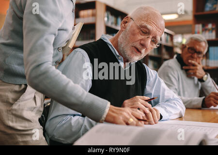 Side view of a lecturer helping a senior man in classroom while another student looks on. Lecturer explaining the concepts from the book of an elderly Stock Photo