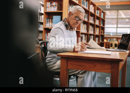 Senior man studying in classroom. Elderly man writing in a book sitting in classroom with a laptop in front. Stock Photo