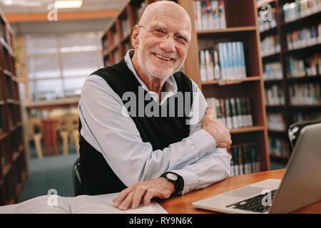 Smiling senior man sitting in a library with his books and laptop on table. Cheerful senior man sitting in library and learning. Stock Photo