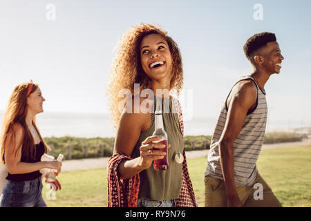 Beautiful young woman with beer walking with friends outdoors on a summer day. Female hanging out with friends outdoors. Stock Photo