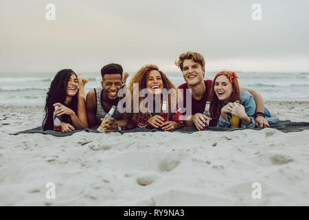 Group of friends lying outdoors on a plaid with beer bottles. Young men and women having beers at the sea shore. Stock Photo