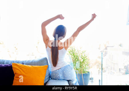 Rear view shot of young woman sitting on couch and stretching after after waking up in the early morning. Stock Photo