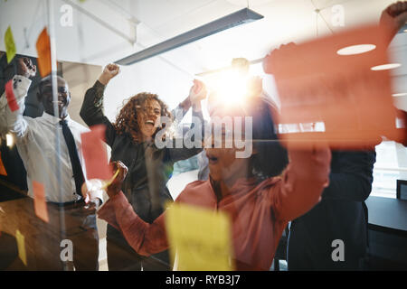 Diverse group of ecstatic businesspeople cheering and jumping up and down while celebrating during a brainstorming session with sticky notes in an off Stock Photo