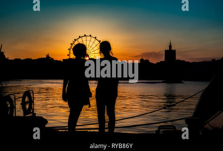 silhouettes of two young girls with ferris wheel in the back at sunset in Malaga port, Spain Stock Photo