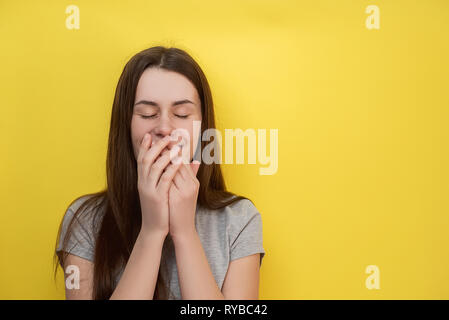 Horizontal shot of good looking young girl giggles joyfully, covers mouth as tries stop laughing, wears gray t-shirt isolated on yellow wall. People a Stock Photo