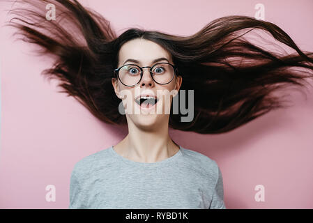Young girl in glasses with flying hair in panic looks with surprised expression, feels nervous in stressful situation, wears gray t shirt, opens mouth Stock Photo