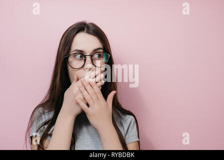 Stunned crazy girl stares with scared expression, covers mouth with both hands, wears gray T-shirt, poses against pink background. People and emotion  Stock Photo