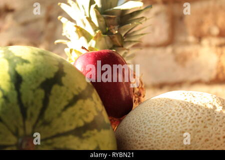 close up de bodegón de frutas en donde aparecen una sandia, un melón, una manzana y una piña y como fondo difuminado una pared de ladrillos Stock Photo