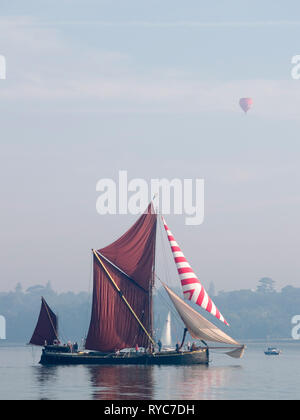 A restored historical sailing barge in the estuary in Suffolk with a hot air balloon passing over head.