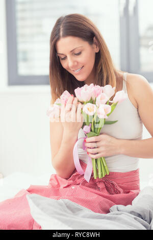 Beautiful attractive woman sitting on the bed holding tenderly in hands a bouquet of spring tulips. Stock Photo