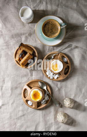 Breakfast with cup of coffee and soft boiled eggs, served in wooden egg cup with salt, pepper and toasted bread, jug of cream over linen tablecloth. F Stock Photo