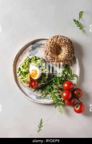 Vegetarian whole grain bagel sandwich with chopped avocado, cream cheese, sun dried tomatoes, egg, arugula served on ceramic plate over white marble b Stock Photo