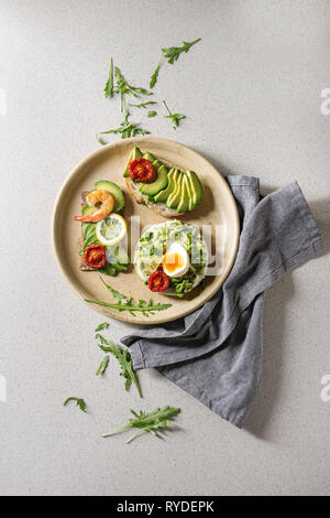 Variety of vegetarian sandwiches with sliced avocado, sun dried tomatoes, egg, shrimps, arugula served on ceramic plate over white grey spotted backgr Stock Photo