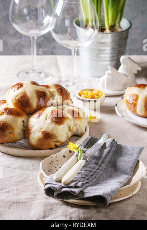 Easter table setting with plate of hot cross buns, narcissus flowers, wine glass on table with linen tablecloth. Stock Photo