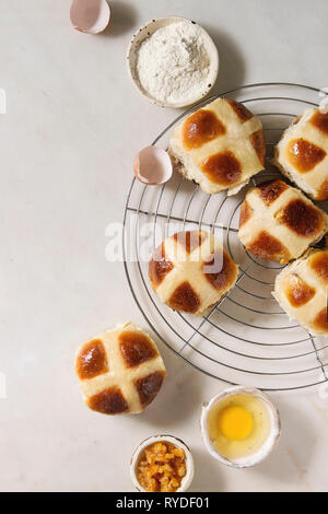 Homemade Easter traditional hot cross buns on cooling rack with ingredients above over white marble background. Top view, space Stock Photo