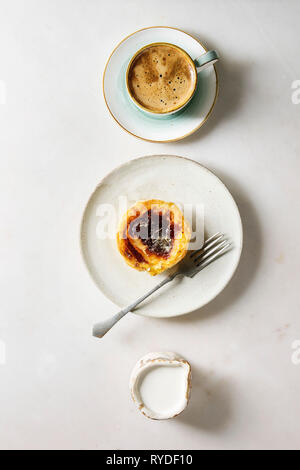 Traditional Portuguese egg tart dessert Pasteis Pastel de nata on ceramic plate with fork, cup of black coffee and jug of cream in row over white marb Stock Photo