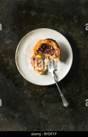 Broken Traditional Portuguese egg tart dessert Pasteis Pastel de nata on ceramic plate with fork over dark metal background. Flat lay, space Stock Photo