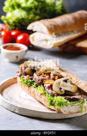 Beef baguette sandwich with champignon mushrooms, green salad, fried onion served on ceramic plate with ingredients above over grey blue table. Close Stock Photo