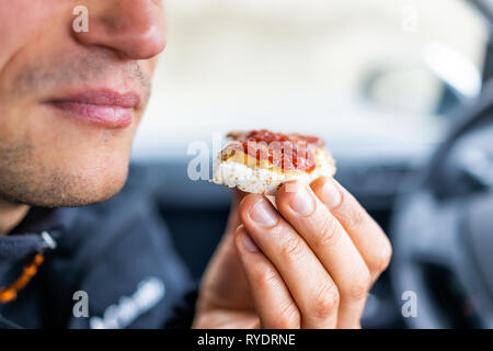 Macro closeup of man hand holding one rice cake by mouth in car road trip blurry background topped with peanut butter, strawberry jam as vegan dessert Stock Photo