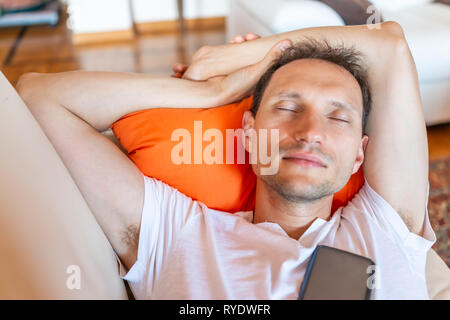 Young man sleeping eyes closed on sofa with smartphone phone relaxing on couch taking a nap during day on orange pillow and happy face Stock Photo