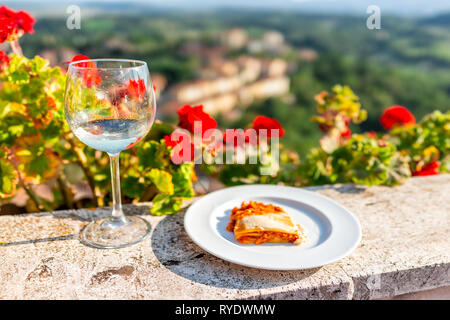 Closeup of vegetarian vegan lasagna slice on plate and white wine on balcony terrace by red geranium flowers outside in Italy with mountain view in Ch Stock Photo