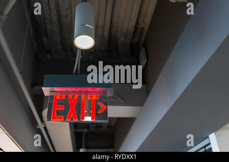Red exit sign hanging from ceiling looking up view with arrow direction and color illuminated fixture in office building closeup Stock Photo