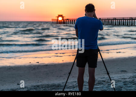 Naples, Florida sunset in gulf of Mexico with sun setting inside Pier bokeh background and back of professional man photographer videographer taking p Stock Photo