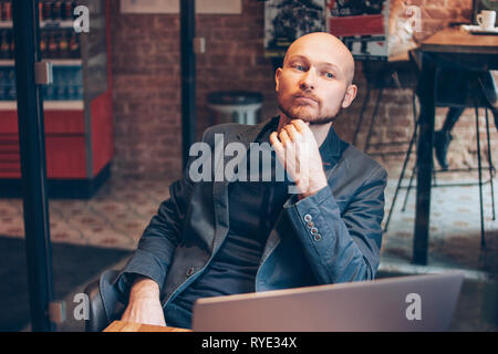 Thinking attractive adult successful bald bearded man in suit with laptop in cafe  Stock Photo