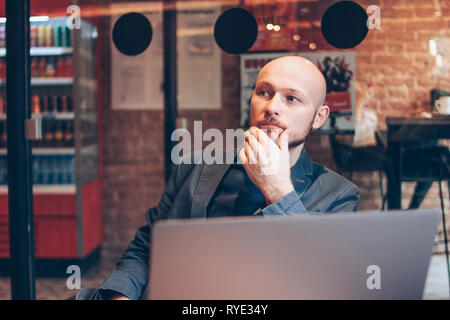 Thinking attractive adult successful bald bearded man in suit with laptop in cafe  Stock Photo