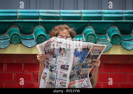 Old Singaporean woman reading newspaper on street at Chinese temple - Singapore Stock Photo