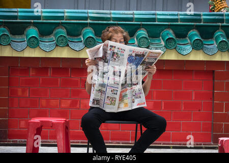 Old Singaporean woman reading newspaper on chair at Chinese temple - Singapore Stock Photo