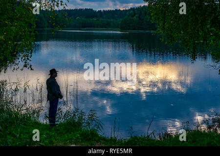 Man in hat is silhouetted as he stands by the side of the water near sunset in the lake district of northeastern Poland near Suwalki Stock Photo