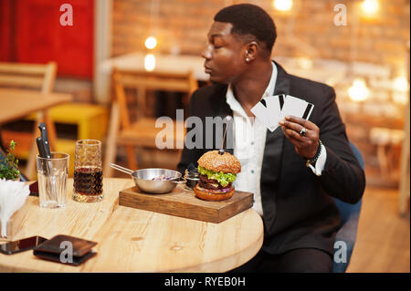 Respectable young african american man in black suit sitting in restaurant hold a lot of credit cards with tasty double burger and soda drink. Stock Photo