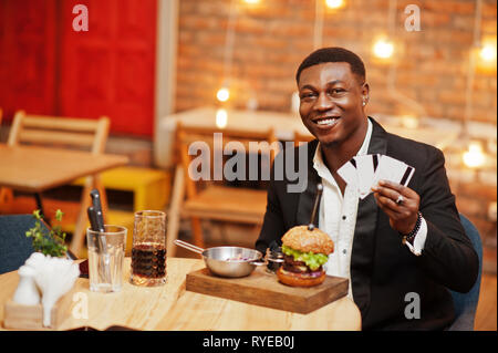Respectable young african american man in black suit sitting in restaurant hold a lot of credit cards with tasty double burger and soda drink. Stock Photo