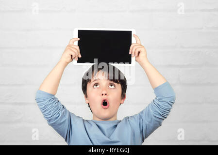 surprised child holding a tablet over his head against brick background Stock Photo
