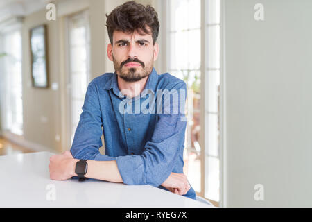Young man wearing casual shirt sitting on white table skeptic and nervous, disapproving expression on face with crossed arms. Negative person. Stock Photo