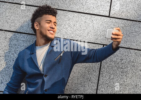 Freestyle. Mulatto guy standing isolated on wall taking selfie on smartphone smiling toothy side view Stock Photo