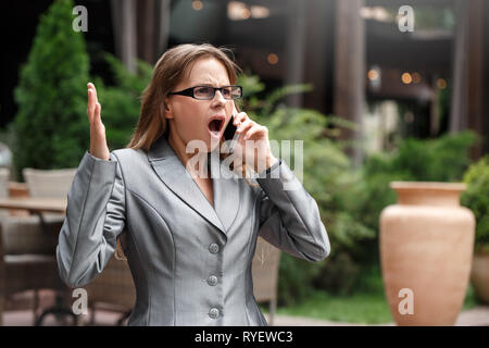 Business woman in eyeglasses standing on the city street having conversation on smartphone hands up shocked Stock Photo