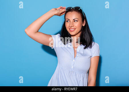 Freestyle. Woman in dress and sunglasses standing isolated on blue wall looking aside joyful Stock Photo