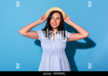 Freestyle. Woman in dress and hat standing isolated on blue wall looking aside dreamful Stock Photo
