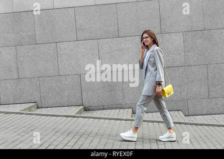 Business casual. Young lady in eyeglasses walking on the city street talking on smartphone happy full body shot Stock Photo