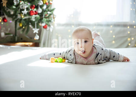 Cute baby girl lying down on bed at home. Christmas tree in background. Girl looks surprised and emotional. Stock Photo