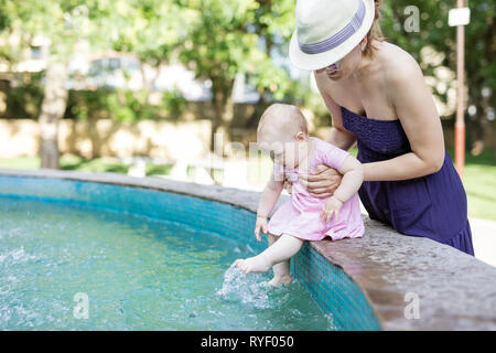 Caucasian young woman holding baby daughter while she is sitting on side of fountain and dangling legs in park Stock Photo