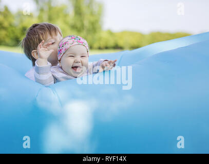 Siblings playing on inflatable sofa outdoors. Baby girl and older brother having fun together in park. Stock Photo