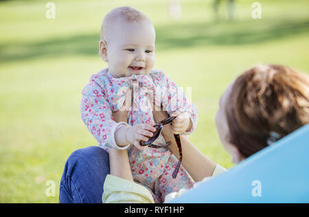 Mother and baby daughter in summer park. Girl is playing with mother's sunglasses while young woman is sitting on inflatable sofa. Stock Photo