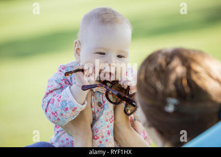 Mother and baby daughter in summer park. Girl is playing with mother's sunglasses while young woman is sitting on inflatable sofa. Stock Photo