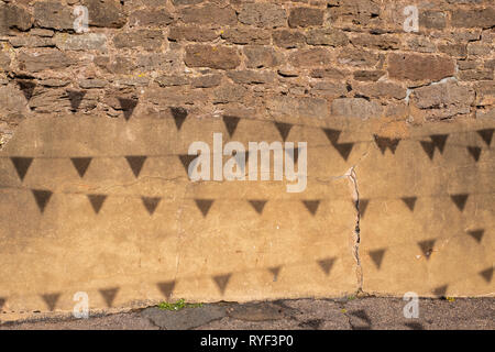 The bright sun creates shadows from bunting hanging overhead create a pattern on a crumbling brick wall. Stock Photo