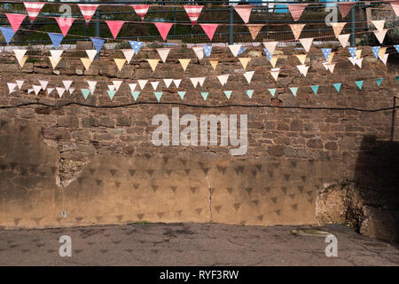 The bright sun creates shadows from tired and worn looking bunting hanging overhead creating a pattern on a crumbling brick wall. Stock Photo