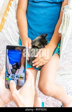 Child sitting in swing chair holding napping kitten in arms and lap cuddling it while mother is taking snap shot photograph image with smartphone Stock Photo