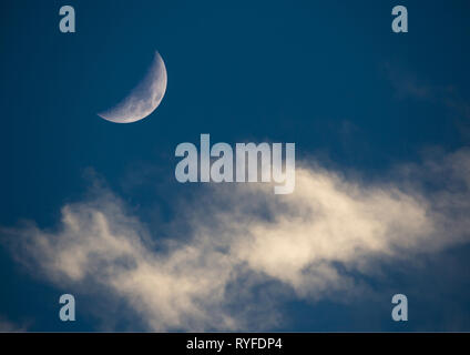 A setting sun illuminates a crescent moon and wispy clouds on a dark blue background. Stock Photo